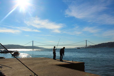 Men fishing at harbor with bosphorus bridge in background against sky