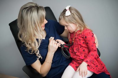 Girl with female doctor holding stethoscope on chair at clinic