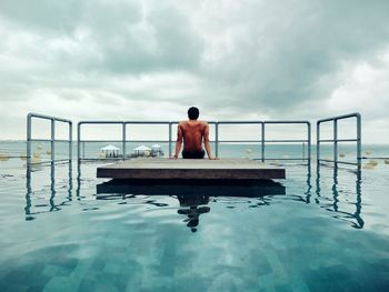 Shirtless man sitting on floating platform in swimming pool against sky