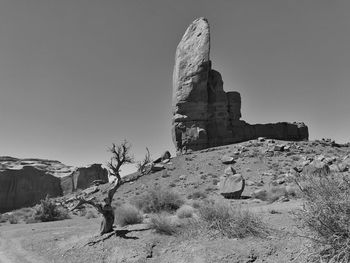 Black and white landscape of a bare tree and tall rounded rock formation in monument valley