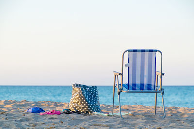 Folding chair by bag on sand at beach against clear sky