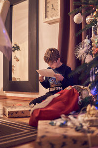Boy reading message while sitting by christmas tree