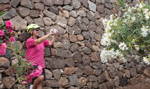 Senior woman photographing flowering plant while standing against stone wall