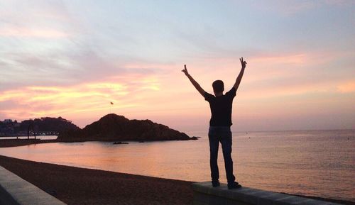 Rear view full length of man showing peace sign at beach against sky during sunset