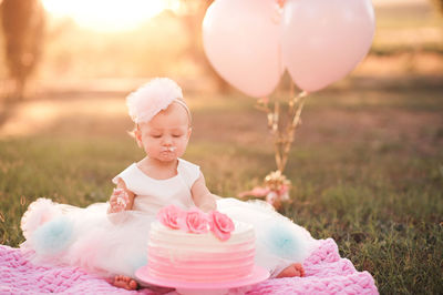 Baby girl 1 year old eating creamy birthday cake sitting on green grass with pink balloons in meadow