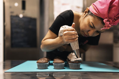 Hispanic female baker in headscarf using pastry bag to squeeze cream on top of chocolate cupcakes while working in bakehouse
