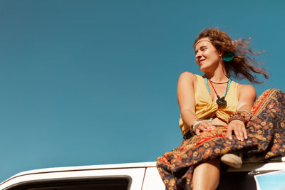 Portrait of young woman sitting on car against clear sky