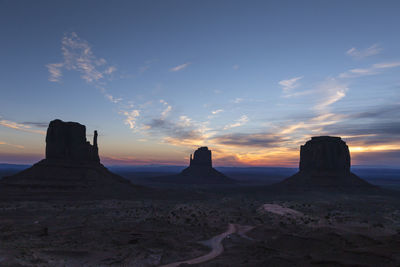 Dirt road leading to three backlit rock monuments at sunrise at monument valley navajo tribal park