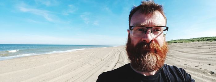 Panoramic view of bearded man wearing eyeglasses at beach against sky during sunny day