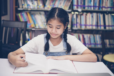 Girl reading book while sitting at table in library
