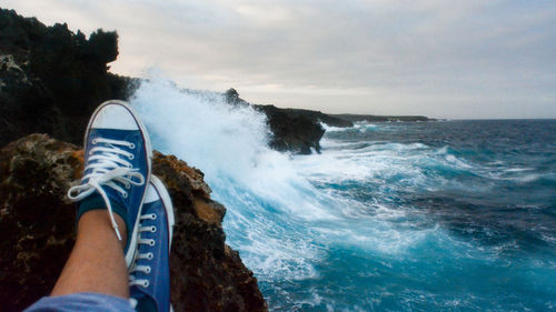 Low section of man sitting on rock by sea against sky