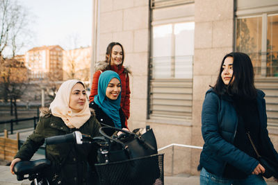 Happy multi-ethnic female friends walking with bicycle against building in city