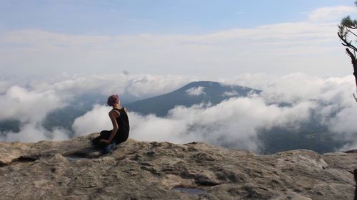 Woman sitting on cliff against mountains amidst clouds
