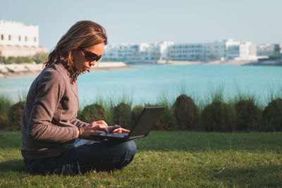 Woman working on laptop while sitting at grassy field against lake