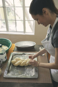 Smiling young woman preparing challah bread from dough in tray on table at home
