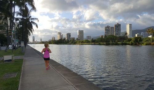 Rear view of woman running alongside canal in city against sky