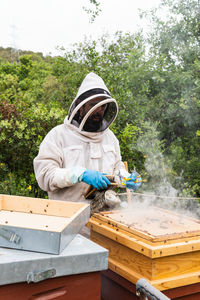 Male beekeeper in protective uniform fumigating beehive with bee smoker while working in apiary