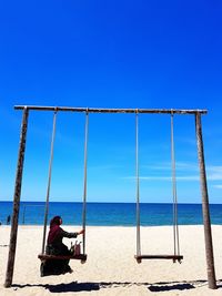Woman in traditional clothing sitting on swing at beach against blue sky during sunny day