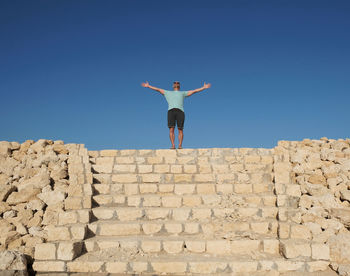 Full length of man with arms outstretched standing on stone wall against clear sky