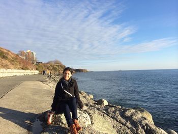 Full length portrait of man sitting on rock by sea against sky