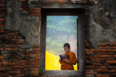 Boy wearing traditional clothing reading book seen through window