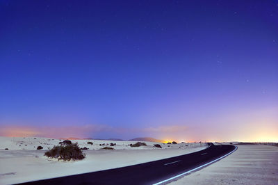 Road amidst snowy field against clear blue sky at dusk