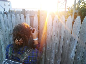 Rear view of woman photographing through wooden fence during sunset