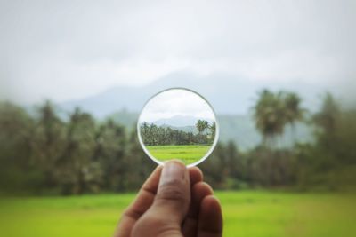 Close-up of hand holding transparent glass on field against sky