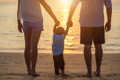 Low section of parents holding son hand while standing at beach during sunset