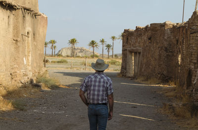 Rear view of adult man in cowboy hat and shirt against abandoned building