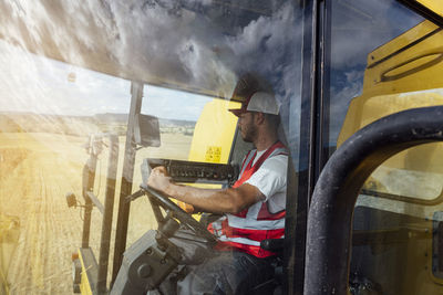 Through window side view of combine operator in uniform harvesting grain crops in agricultural field while working in rural area