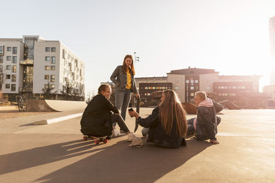 Friends looking at teenager showing mobile phone to girl sitting on skateboard in city