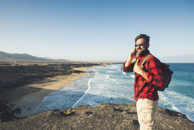 Bearded mature man with backpack standing at beach against sky