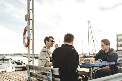 Men sitting on table at harbor against sky