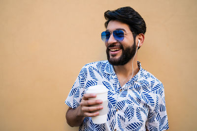 Smiling young man holding coffee cup sanding by wall outdoors