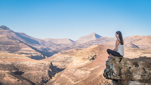 Side view of woman sitting on cliff by mountains against clear sky