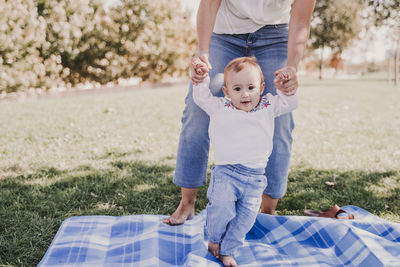Low section of mother holding baby girl hands while standing on blanket