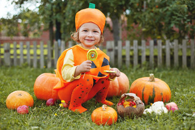 Full length of cute girl wearing costume sitting by pumpkin at park