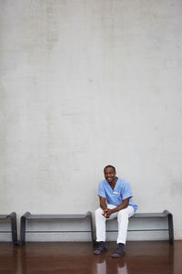 Full length of confident mid adult male nurse sitting against wall at hospital