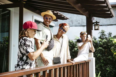 Smiling parents looking at daughter using smart phone standing by son on porch of villa