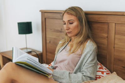 Friendly charming blonde woman reading book on bed in bright interior