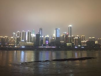 Illuminated buildings by river against sky at night