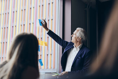 Confident mature manager sticking adhesive notes on window in board room during meeting at creative office