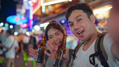 Portrait of smiling young couple holding beer bottles while standing on street in city at night