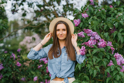 Charming young woman in a denim shirt and a straw hat walks among the blooming lilacs in the spring