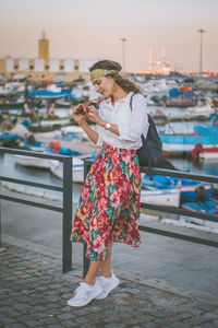 Young woman looking at sunglasses while standing by railing during sunset