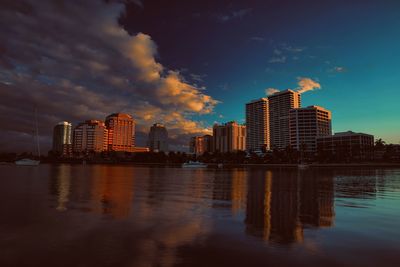 Illuminated buildings by river against sky at sunset