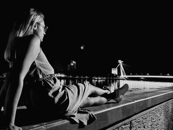 Side view of woman sitting on wall at the river at night