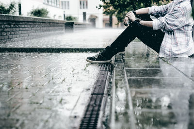 Low section of young man sitting on footpath during rain