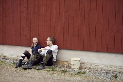 Full length of siblings talking while sitting against barn